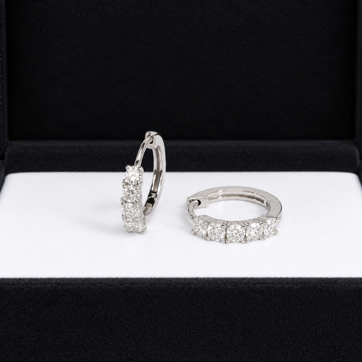 A pair of silver huggie hoop earrings set with clear cubic zirconia stones, displayed inside an open black jewelry box with a white insert.