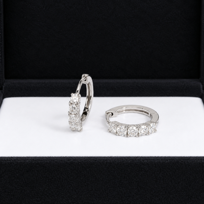 A pair of silver huggie hoop earrings set with clear cubic zirconia stones, displayed inside an open black jewelry box with a white insert.