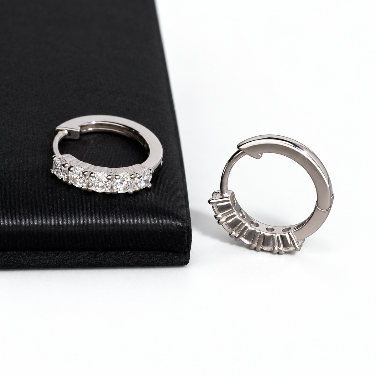 A pair of silver huggie hoop earrings with clear baguette-cut stones, shown on a white background with one earring resting on a black textured surface.