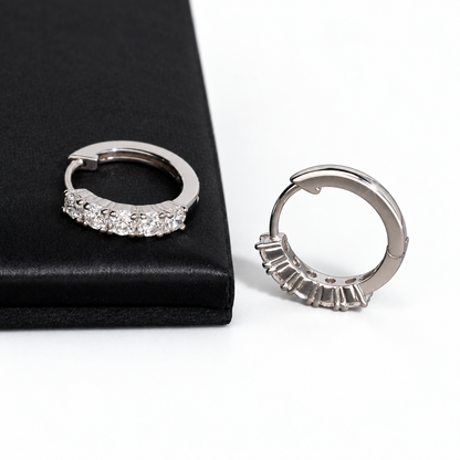A pair of silver huggie hoop earrings with clear baguette-cut stones, shown on a white background with one earring resting on a black textured surface.