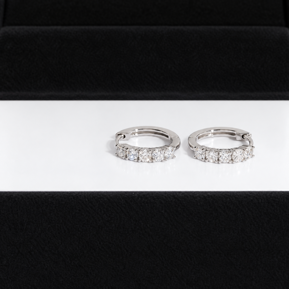 A pair of silver-toned huggie hoop earrings set with clear pavé diamonds, displayed on a white surface with a black velvet jewelry box in the background.