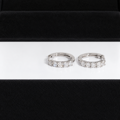 A pair of silver-toned huggie hoop earrings set with clear pavé diamonds, displayed on a white surface with a black velvet jewelry box in the background.