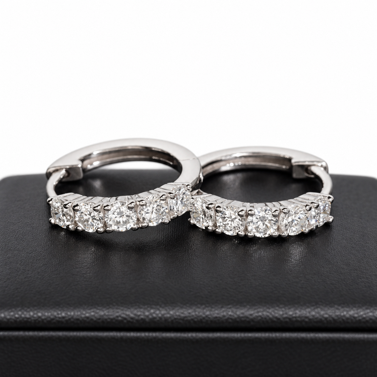 A pair of silver hoop earrings set with round-cut clear gemstones, displayed on a black textured jewelry box against a white background.
