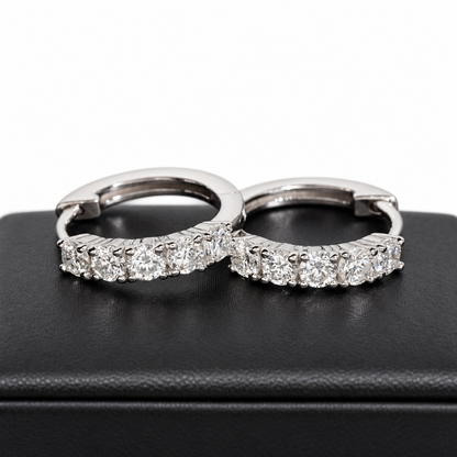 A pair of silver hoop earrings set with round-cut clear gemstones, displayed on a black textured jewelry box against a white background.
