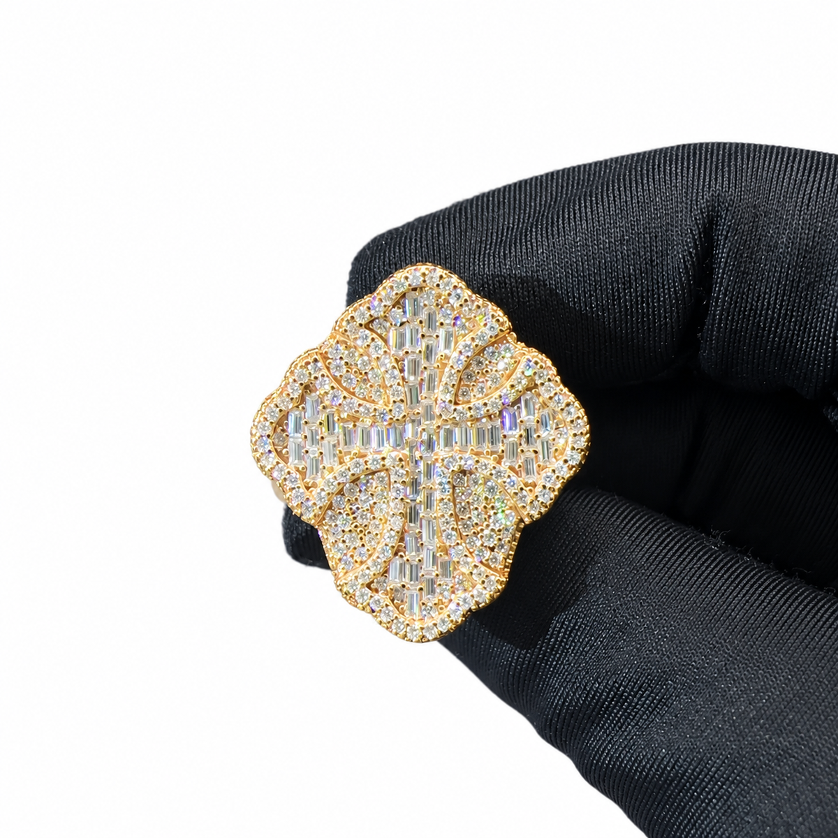 A gold-toned clover-shaped brooch or pin, heavily embellished with clear crystals, held by a hand wearing a black glove against a plain white background.