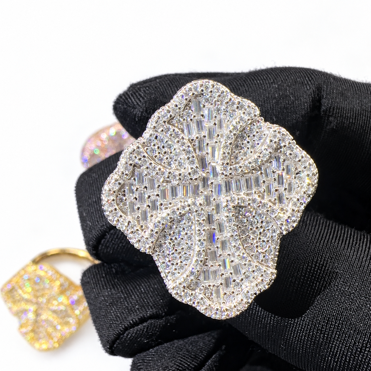 A large floral cross ring covered in clear crystals on a silver-tone setting, held on a black gloved hand. A smaller gold-toned version is partially visible in the lower left against a white background.