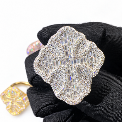 A large floral cross ring covered in clear crystals on a silver-tone setting, held on a black gloved hand. A smaller gold-toned version is partially visible in the lower left against a white background.