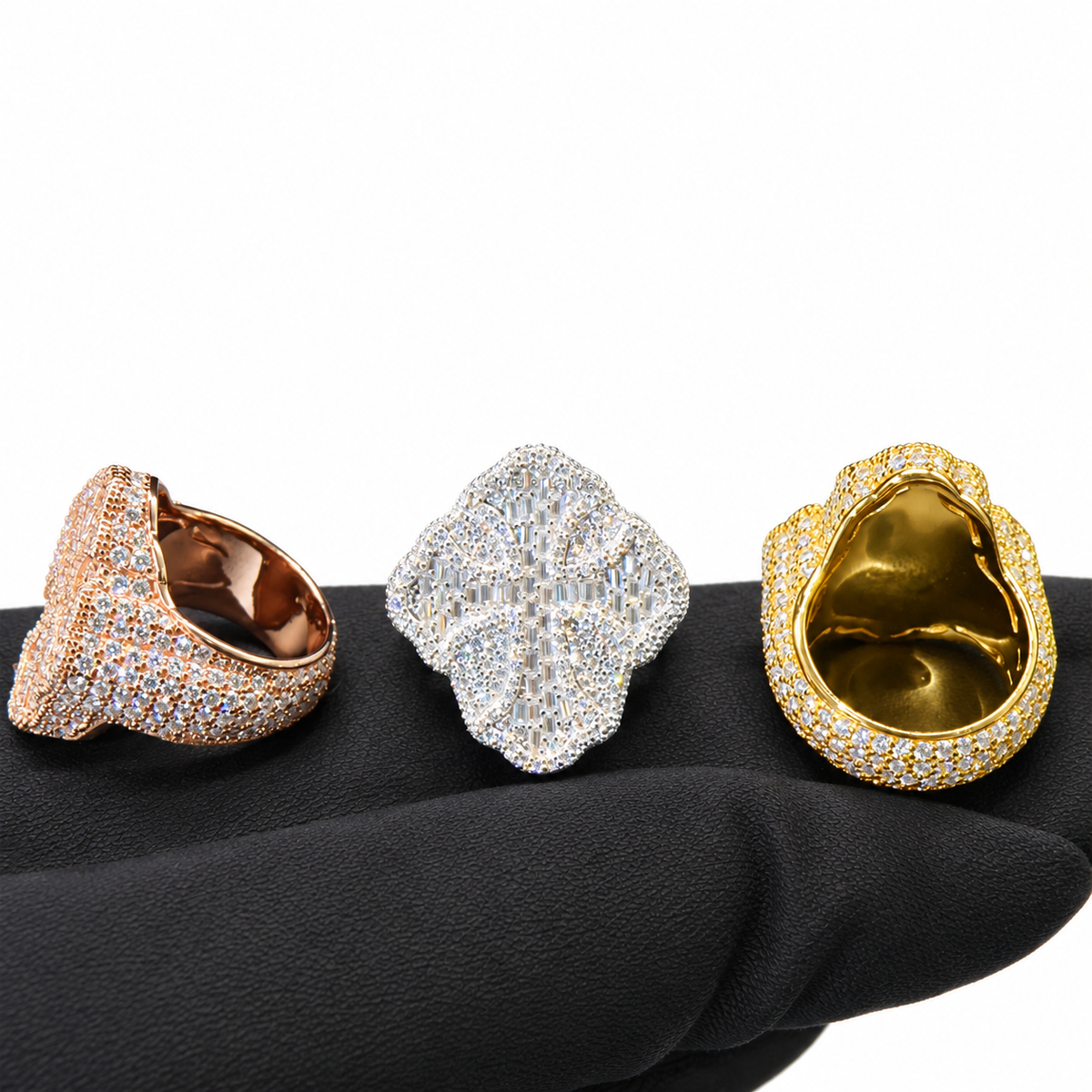 Three oversized statement rings with pavé-set clear stones are displayed on a black textured surface against a white background. The rings are shown in rose gold, silver, and yellow gold tones.