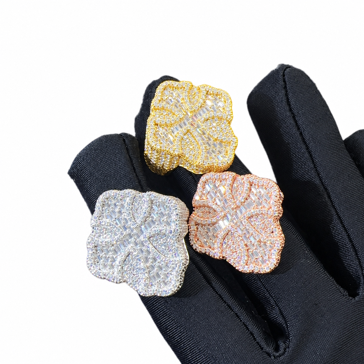 Three flower-shaped pave rings in yellow gold, rose gold, and silver tones, displayed on a black glove against a white background.