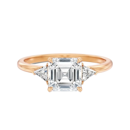 A rose gold engagement ring featuring a central emerald-cut diamond surrounded by two smaller round diamonds, set on a polished rose gold band against a clean white background.
